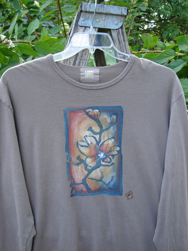 1999 Long Sleeved Vented Tee Orchid Sprig Shale Size 1, featuring a stylized flower panel on the front, hangs on a clear plastic hanger amidst green foliage, showcasing its ribbed neckline and vented sides.