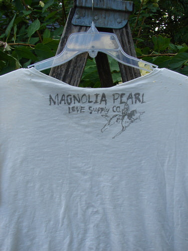 Magnolia Pearl NWT Cotton Europa V Neck Boyfriend Tee, True White OSFA, draped on a hanger outdoors, features stenciled MAGNOLIA PEARL Love Supply Co. on back, highlighting its raw edges and hand distressing.