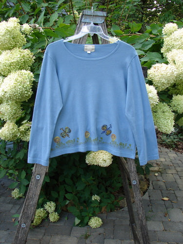 1999 Round Neck Rib Top Butterfly Blue Stone Size 2 displayed on a weathered ladder, showcasing whimsical embroidered flowers, mushrooms, and ladybugs along its hem, in a lush garden setting.