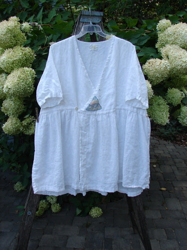 1999 Linen Adjustable Wrap Dress Fish White OSFA: A white V-neck blouse hangs from a wooden ladder, showcasing its adjustable crossover front and empire waistline, surrounded by lush garden foliage and brick paving.