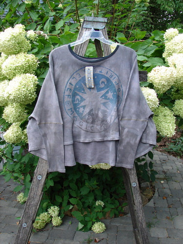 Barclay NWT Thermal Crop Top Belief Magic Mottled Spirit OSFA, featuring a faded circular graphic with themed text, hangs on a clear hanger over a wooden stepladder amid lush foliage and white hydrangeas.