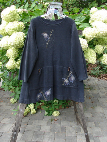 The 1998 Heavy Berber Kindlewood Tunic Leaf Sprig Black OSFA features an A-line cut with toggle buttons, front patch pockets, and delicate embroidery, hanging elegantly against a garden backdrop.
