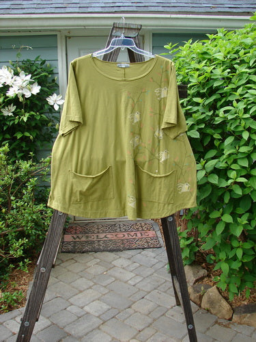 A green Barclay Double Pocket Twinkle Top with a song bird design on it, made from medium weight organic cotton. Features include a rounded banded bottom shape, a feminine neckline, and lower exterior pockets.