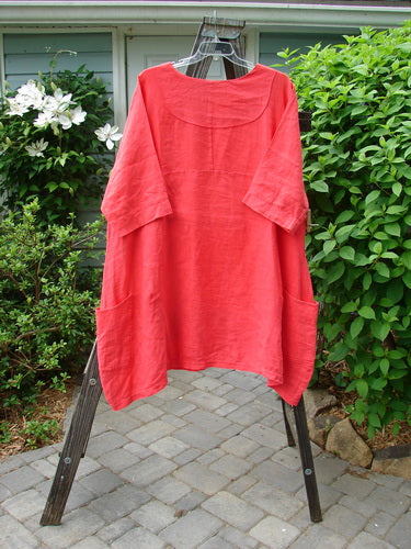 Barclay Linen Cross Over Dress in Geranium, Size 2. A red shirt on a rack, featuring a cross over front V neckline, drop shoulders, and a curved empire waist seam. Varying side lengths and double drop flop side pockets.