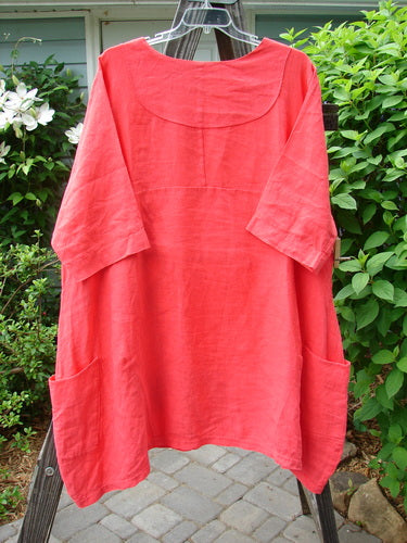 A medium weight linen dress in geranium color, featuring a cross-over front V neckline, drop shoulders, and a downward curved empire waist seam. The dress has varying side lengths, double drop flop side pockets, and is painted in the nature lookout theme. Size 2.