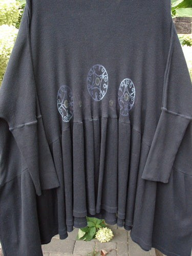 1997 Thermal Bungalow Jacket Celtic Medallion Cast Iron OSFA, featuring a loose black knit design with long sleeves, gathered lower panel, circular motifs, and colorful buttons, hangs outdoors against garden foliage.