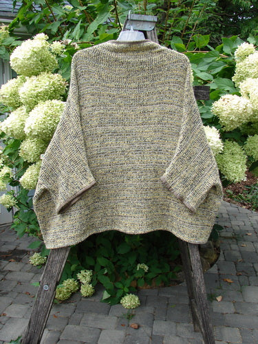 Barclay Alpaca Big Square Mock Collar Sweater Marled Sun OSFA drapes on a hanger, showcasing a textured knit with dolman shoulders and three-quarter sleeves, set against a lush garden backdrop.