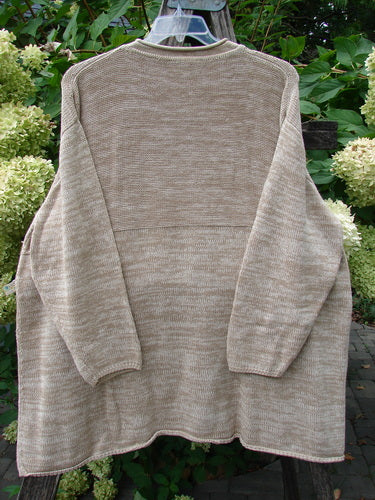 Barclay Jennifer's Rollneck Sweater Marled Aspen OSFA, featuring a hand-knit stockinette-stitch body, raglan sleeves, and an A-line drape, displayed outdoors with a garden backdrop of green leaves and pale blossoms.