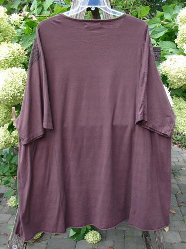 Barclay Art Tee Grid Flower Lower Lily Red Plum OSFA, loose-fitting tunic-length top with drop shoulders and varying hemline, displayed on a hanger against a lush outdoor backdrop.