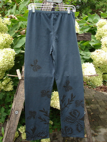 Barclay NWT Cotton Lycra Straight Pant Botanicals Lux Silhouette Size 1, featuring black stenciled butterflies and leaves, elastic waistband, straight-leg cut, displayed on a hanger against a rustic garden backdrop.