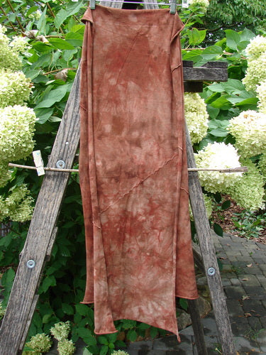 Barclay Diagonal Scarf Wrap Belief Spirit Magic Mottled Rust OSFA draped on a wooden A-frame ladder, showcasing its sweet raw curly edges and exterior diagonal sectional seams against a shrub backdrop.