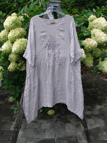 Barclay NWT Linen Wave Pocket Dress in Floral Bunch Vintage Quartz, featuring a loose fit with a wide neckline, elbow-length sleeves, uneven hem, and front pockets, displayed on a wooden ladder in a garden setting.