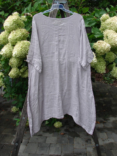Barclay NWT Linen Wave Pocket Dress in Vintage Quartz features a loose-fitting, smock-style design with a rounded neckline, seamless bodice, and pinched sleeves, hanging outdoors against a garden backdrop.