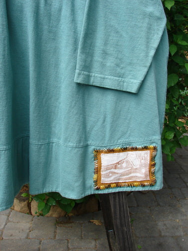1993 PMU Patched Modernismo Cardigan in Grey Green. Close-up of a blue shirt with whip-stitched accents. Kangaroo front pockets, A-lined swing, drawcord back, oversized patches, and banded hemline.