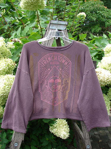 Barclay NWT Fleece Donna Sweatshirt Frenchtown Logo Fig OSFA hanging on a wooden ladder, showcasing its wide, slightly A-lined shape, drop shoulders, and distinctive Frenchtown theme graphic in a garden setting.