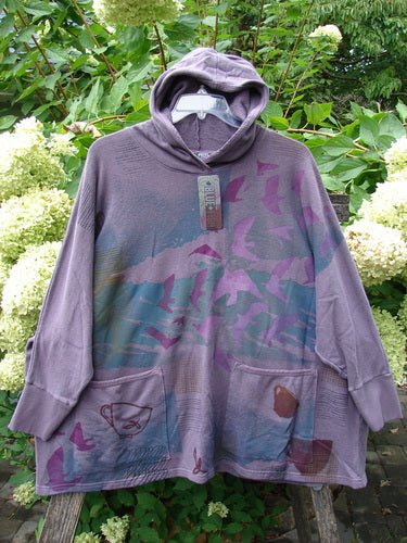 Barclay NWT Cotton Hemp Folklore Hoodie Morning Dove Aura OSFA: Loose-fitting, washed-purple tunic-length hoodie with stylized bird print, kangaroo pockets, and drawstring hood, hanging against white hydrangeas.