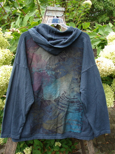 Barclay Cotton Hemp Hip To Be Square Hoodie Rear Botanicals Black OSFA, showcasing a muted abstract print on a blue-gray hooded sweatshirt, draped over a wooden ladder amidst lush greenery and hydrangeas.