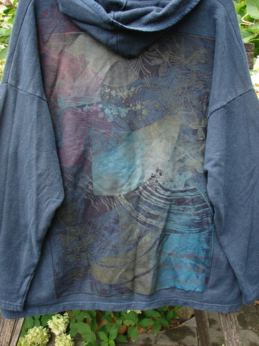 Barclay Cotton Hemp Hip To Be Square Hoodie Rear Botanicals Black OSFA draped over a wooden chair, showcasing its abstract wave-like and botanical print, highlighting its cozy fleece texture and unique design features.