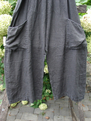 Barclay Linen Crop Two Pocket Pant Unpainted Black Size 2, featuring wide legs, oversized patch pockets, and a full elastic waist, hangs over a bench. The pants exhibit a billowy flair and vertical seams.