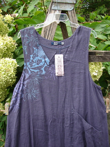 Barclay NWT Linen Explorer Jumper in Purple River Mist by Blue Fish, featuring a sleeveless, slate-blue design with a floral pattern, scoop neckline, and patch pocket, hanging against a wooden garden structure.