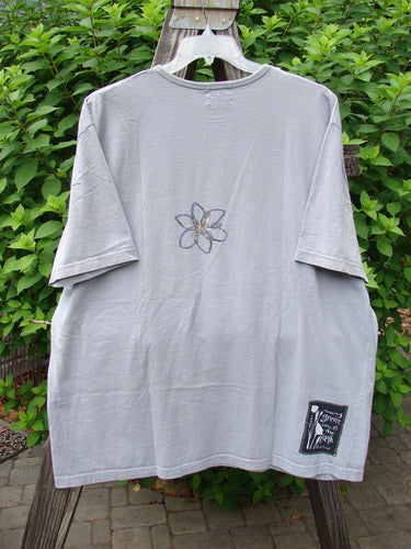 A faded lavender short-sleeved tee with a rolled neckline and drop shoulders. Features a flower made of wire and a leaf detail. Size 2.