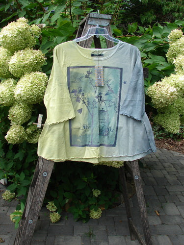 Barclay NWT Three Quarter Sleeved Cafe Tee Free Bird Mottled Violet Citron Size 0, featuring a botanical/birdcage print, scalloped elbow-length sleeves, and original hang tag, displayed on a hanger against a garden backdrop.