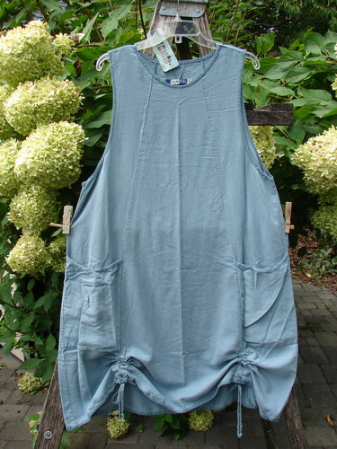 Barclay NWT Lyocell Joyous Jumper Unpainted Sea Green Size 0, a sleeveless tunic with large patch pockets and hem draw-string ties, displayed outdoors against lush foliage and hydrangea blooms.