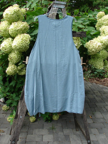 Barclay NWT Lyocell Joyous Jumper Unpainted Sea Green Size 0: A sleeveless, loose-fitting tunic dress with deep arm openings and double front pockets, displayed outdoors on a wooden ladder with floral backdrop.
