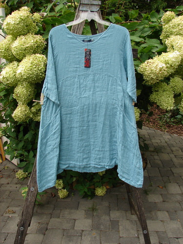 Barclay NWT Linen Zen Pocket Tunic Dress Unpainted Turquoise Size 0 hangs outdoors, showcasing its lightweight, crinkled linen fabric, three-quarter sleeves, A-line shape, and dual drop pockets against hydrangeas.