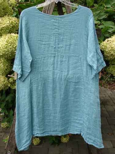 Barclay NWT Linen Zen Pocket Tunic Dress Unpainted Turquoise Size 0 displayed on a hanger, featuring a rounded neckline, three-quarter sleeves, A-line shape, curved waist seam, dual exterior pockets, and varying hemline.