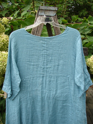 Barclay NWT Linen Zen Pocket Tunic Dress Unpainted Turquoise Size 0, displayed on a hanger in a garden, features an A-line shape, three-quarter sleeves, and dual pockets, highlighting its relaxed design.