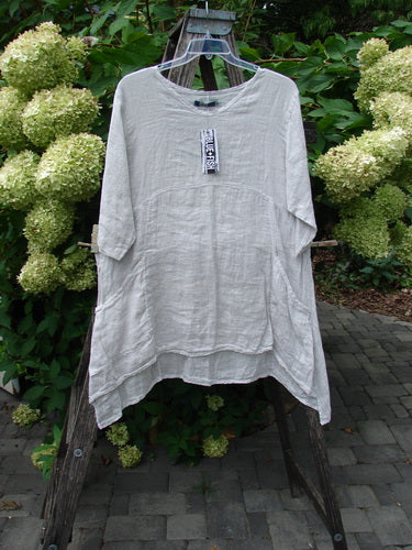 Barclay NWT Linen Zen Pocket Tunic Unpainted Dove Size 0 displayed on a hanger, featuring a loose fit, three-quarter sleeves, oval neckline, dual pockets, and curved hemline against hydrangea blooms.