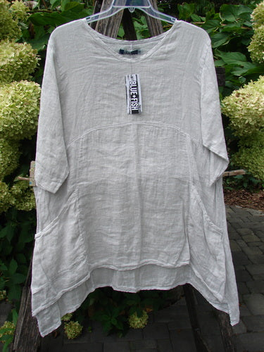 Barclay NWT Linen Zen Pocket Tunic Unpainted Dove Size 0: Loose-fitting, natural-beige linen tunic with a rounded neckline, three-quarter sleeves, subtle bust seaming, side pockets, and an uneven hem, hung on a clear plastic hanger.