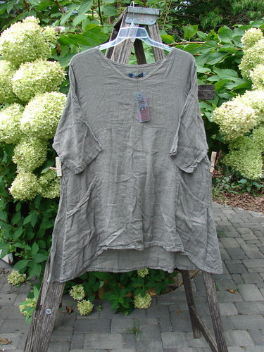 Barclay NWT Linen Zen Pocket Tunic Unpainted Stone Beach Size 0 displayed on a wooden ladder, showcasing its loose, A-line fit, roll-tab sleeves, dual patch pockets, and attached tag against a patio backdrop.