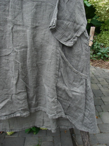 Barclay NWT Linen Zen Pocket Tunic Unpainted Stone Beach Size 0 hangs outdoors, showcasing its grey, wrinkled linen fabric, large curved side pocket, short sleeve, and A-line shape with three-quarter sleeves.