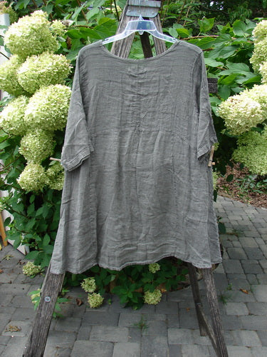 Barclay NWT Linen Zen Pocket Tunic Unpainted Stone Beach Size 0, displayed on a ladder, features a loose-fit, gray-green linen tunic with three-quarter sleeves, dual pockets, and a varying hemline.