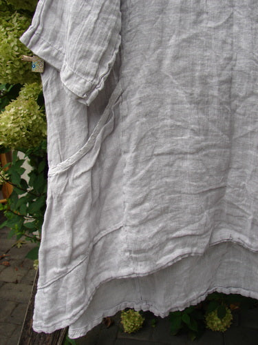 Barclay NWT Linen Zen Pocket Tunic Unpainted Pearl Smoke Size 0, hanging outdoors, features a light-gray, loose-weave linen with three-quarter sleeves, front pocket, and varying hemline, set against lush greenery.