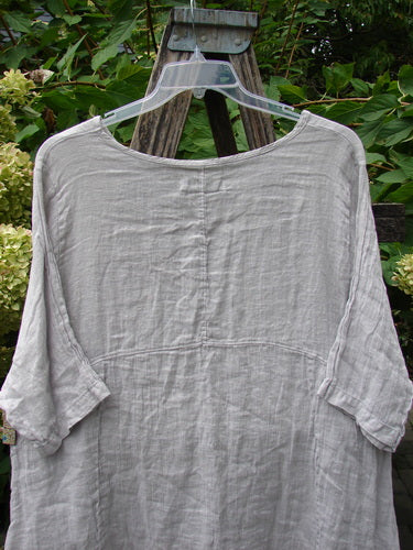 Barclay NWT Linen Zen Pocket Tunic Unpainted Pearl Smoke Size 0: Loose-fitting, light gray tunic with three-quarter sleeves, displayed outdoors on a hanger against wooden trellis, featuring an A-line shape and dual exterior pockets.