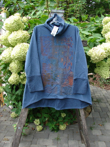 Barclay NWT Fleece Cowl Neck Sweatshirt Metallic Daisy Vintage Indigo Size 0 hangs on a wooden ladder, showcasing its abstract pattern in rusty-orange and turquoise. The cowl neck with attached tag is prominently visible.