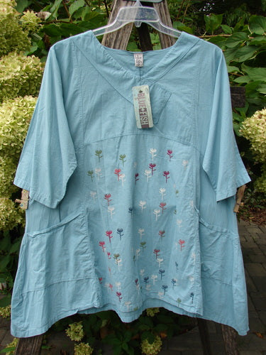 Barclay NWT Batiste Cross Over Urchin Pocket Tunic Tiny Tulip Turquoise Size 0, featuring embroidered flowers, V-neck, elbow-length sleeves, and hip-level pockets, displayed on a plastic hanger against foliage.
