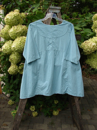 Barclay NWT Batiste Cross Over Urchin Pocket Tunic Tiny Tulip Turquoise Size 0, displayed on a wooden frame outdoors with a lush, floral backdrop, highlighting its airy design and unique sectional panels.