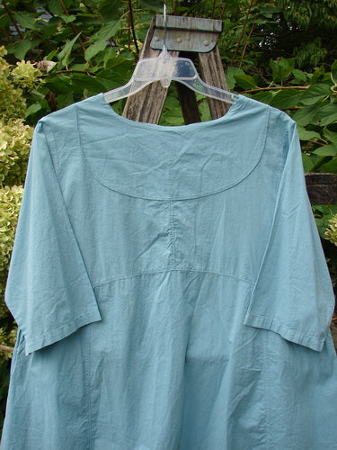 Barclay NWT Batiste Cross Over Urchin Pocket Tunic Tiny Tulip Turquoise Size 0, featuring three-quarter sleeves, a crossover neckline, and exterior flop pockets, displayed on a clear hanger against green foliage.