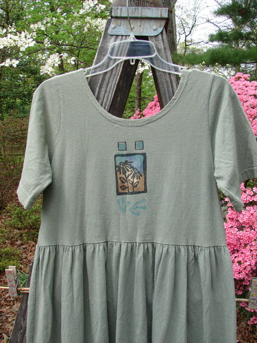 1996 Short Sleeved Simple Dress Sunflower Cricket Size 0 displayed on a hanger, featuring a rounded neckline, drop empire waist seam, and full sweeping skirt with a sunflower garden theme painting.