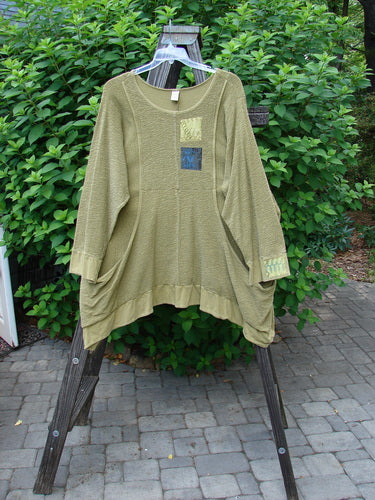 A Barclay Patched Crepe Perennial Tunic Top in Woods Olive, size 2, displayed on a rack. The tunic features textured cotton crepe fabric, drop wrap side pockets, and a curved empire waist seam. It has S-shaped seams, a varying dip hemline, and banded lower sleeves and hemline. The tunic also has oversized patches, a rounded neckline, and a drawcord at the rear.