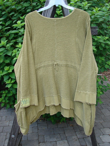 A green shirt on a rack, part of the Barclay Patched Crepe Perennial Tunic Top collection. Made from thick mixed textured cotton crepe, it features a weighted stretch fabric, wrap side pockets, an empire waist seam, S-shaped seams, a varying dip hemline, banded lower sleeves and hemline, cozy lower cuffs, and a handy drawcord rear. Superior double oversized patches and a rounded neckline complete the design.