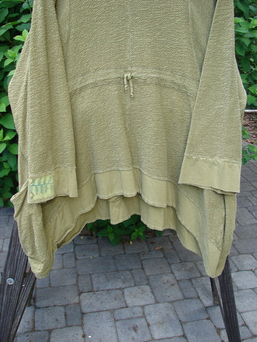 A Barclay Patched Crepe Perennial Tunic Top in Woods Olive, size 2. Textured cotton fabric with wrap side pockets, empire waist seam, S-shaped seams, varying dip hemline, banded sleeves and hemline, cozy cuffs, and a drawcord rear. Double oversized patches and rounded neckline.