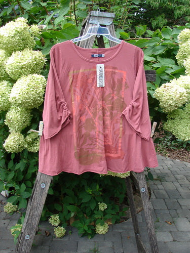 Barclay NWT Draw Gather Sleeve Cafe Top Free Bird Desert Size 0 displayed on a wooden ladder, features 3/4-length ruched sleeves, rounded neckline, and subtle graphic, surrounded by white hydrangeas, showcasing its unique design.