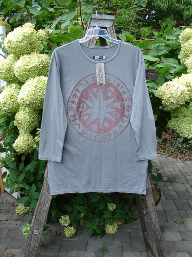 Barclay NWT Long Sleeved Studio Tee in Grey Moonlight, featuring a faded red compass graphic and Belief Spirit Magic theme, displayed on a wooden ladder against hydrangeas.