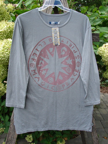 Barclay NWT Long Sleeved Studio Tee Belief Spirit Magic Grey Moonlight Size 0, featuring a circular faded-red graphic with words and stars, hangs on a clear hanger against a rustic outdoor backdrop.