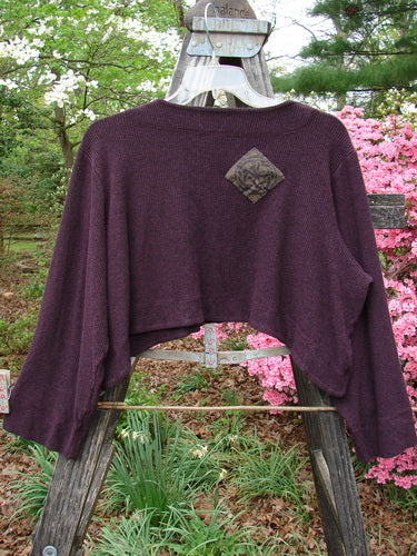 Purple 2000 Patched Rayon Lycra Rib Overtop in Cassis Heather displayed on a wooden swing, showcasing a dramatic upward scooping hemline and Celtic-themed painted patches, highlighting its unique texture and stitchery.